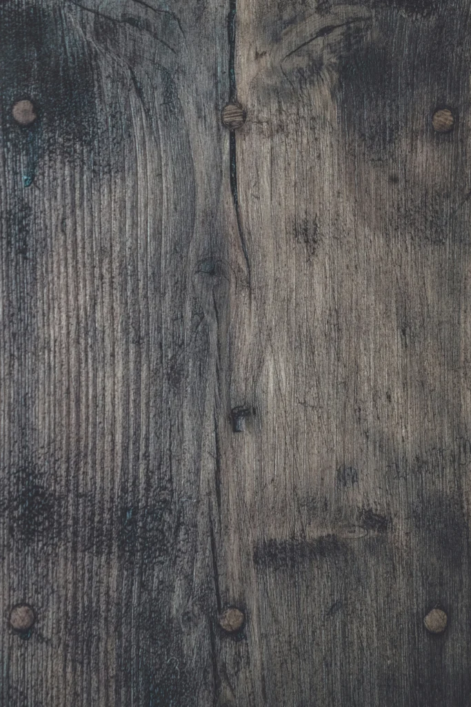 Close-up of a weathered wooden surface with visible knots, grain patterns, and a vertical crack. Five metal studs are embedded, adding a rustic texture.