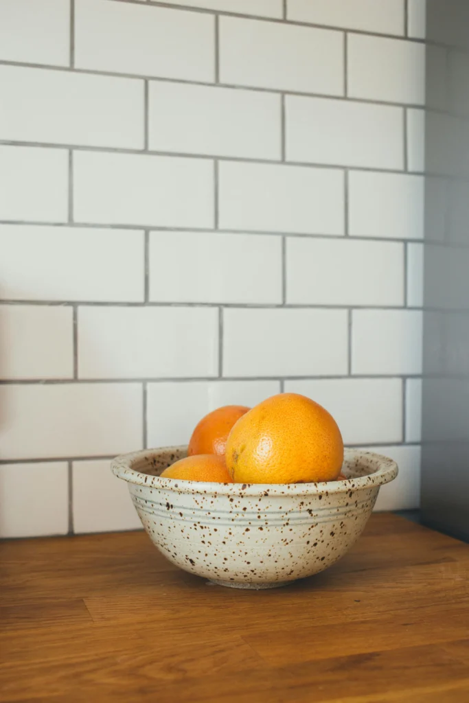 A speckled ceramic bowl with three oranges is placed on a wooden counter against a white tiled wall.