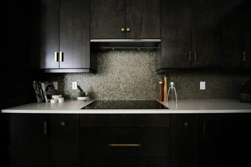 Modern kitchen with dark cabinets, a white countertop, and a textured backsplash. Induction cooktop, wooden pepper mill, and clear glass bottle are visible.