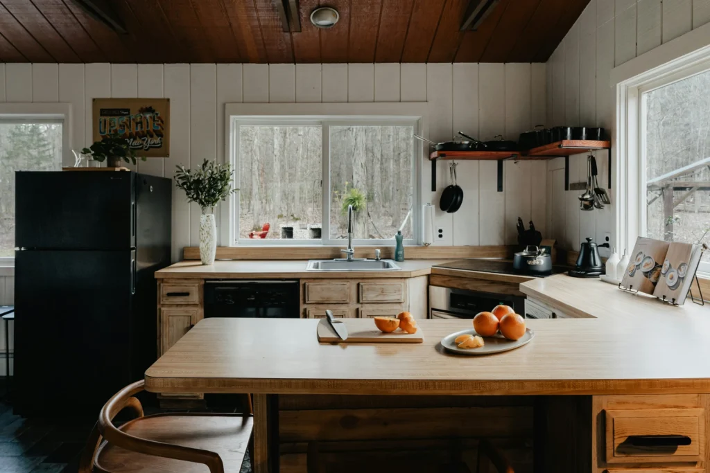 Cozy kitchen with wooden cabinets, black appliances, a large window, and a countertop holding oranges and kitchenware. Natural light illuminates the space.