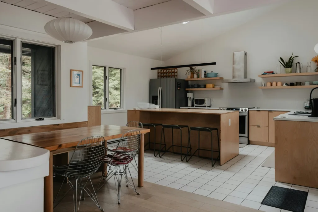 Modern kitchen with wooden cabinets, an island with stools, dining table with wire chairs, and natural light from large windows.