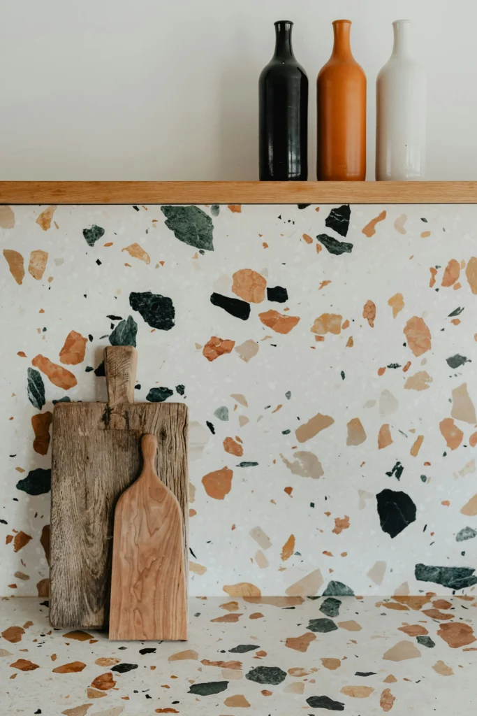 Terrazzo countertop with two wooden cutting boards leaning against it, and three decorative bottles on a wooden shelf above.