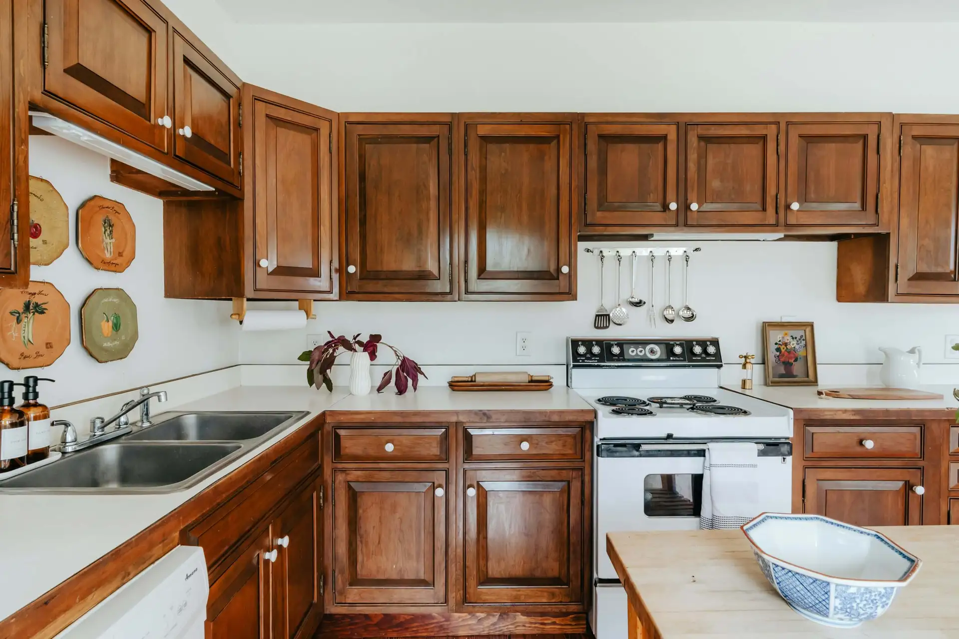 A kitchen with wooden cabinets, a white countertop, a stove with hanging utensils, a double sink, and decorative plates on the wall. A small plant and a bowl are on the counter.
