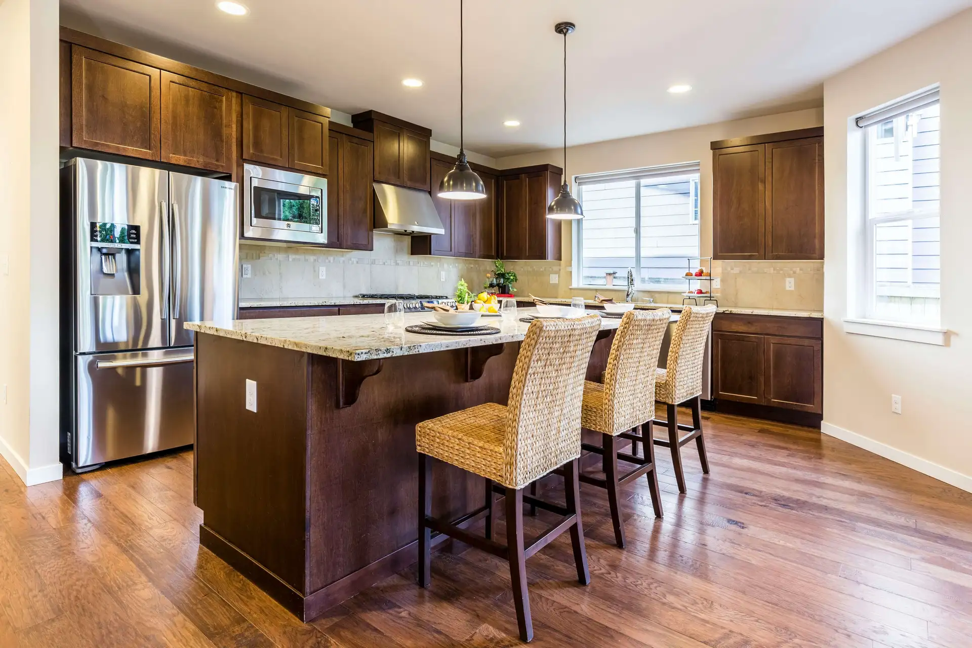 Modern kitchen with stainless steel appliances, wooden cabinets, and an island with wicker barstools. Bright lighting and hardwood flooring complete the room.