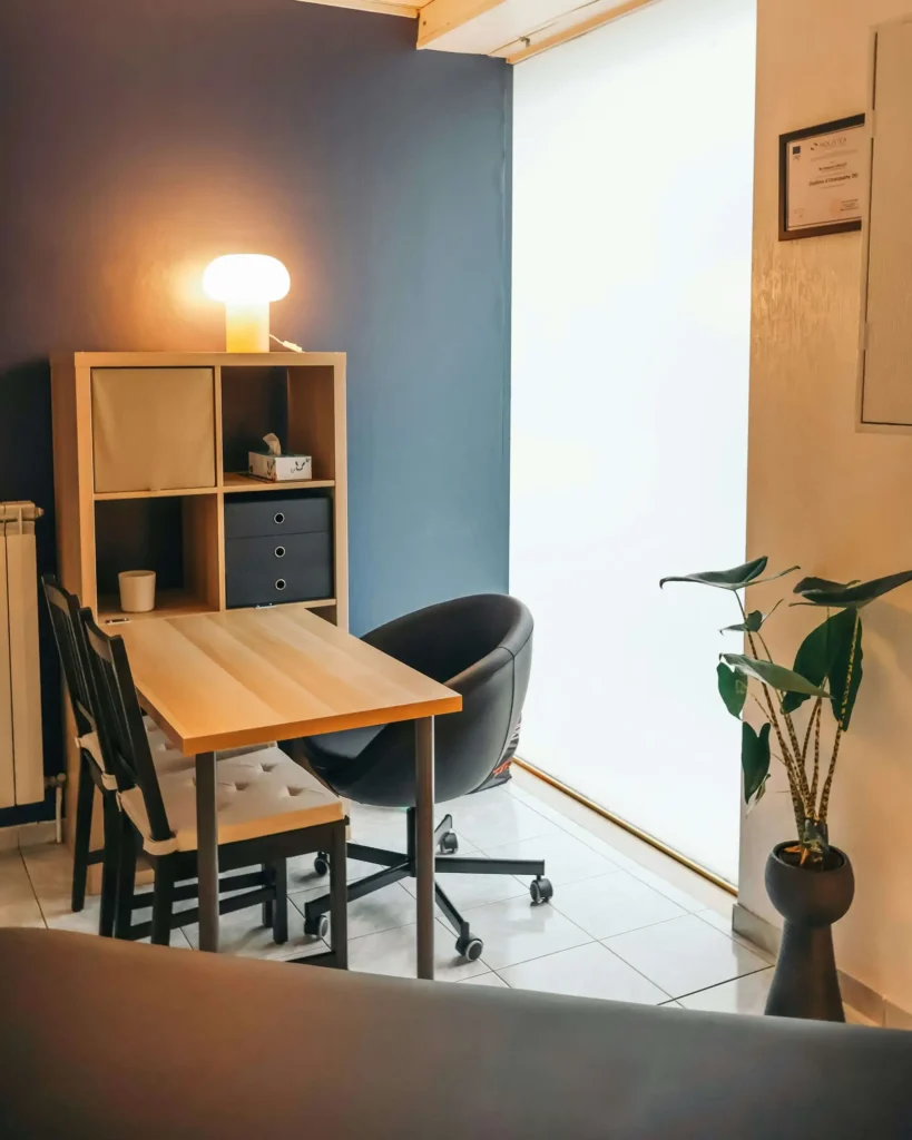 Office corner with a wooden table, three chairs, a shelf with storage boxes, a potted plant, and a lamp. A framed certificate hangs on the blue wall. Natural light from a large window.