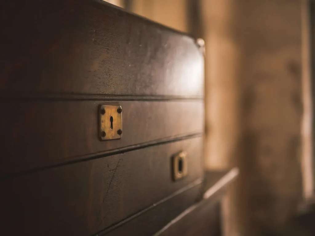 Close-up of an old wooden chest, showing a brass keyhole on its front. The background is softly blurred, highlighting the chest's dark, polished wood and metal detail.