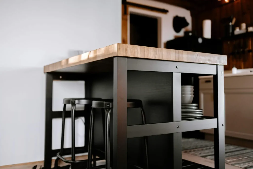 A wooden kitchen island with two metal stools underneath and a stack of white bowls on the shelf, set in a modern kitchen.