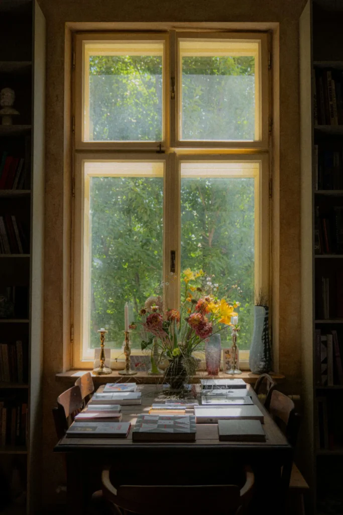 A table with scattered books and a vase of flowers sits in front of a window, through which trees are visible.