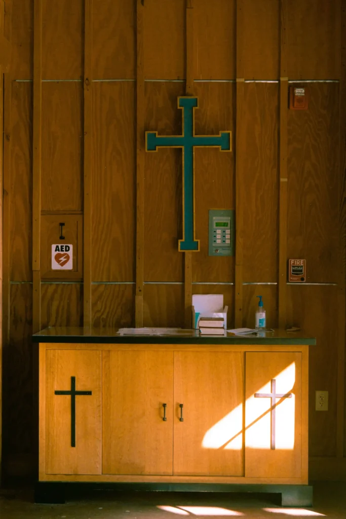 A wooden altar with a large blue-outlined cross above it and a smaller cross on the front. A stack of books and hand sanitizer sit on the altar. An AED and fire extinguisher sign are on the wall.