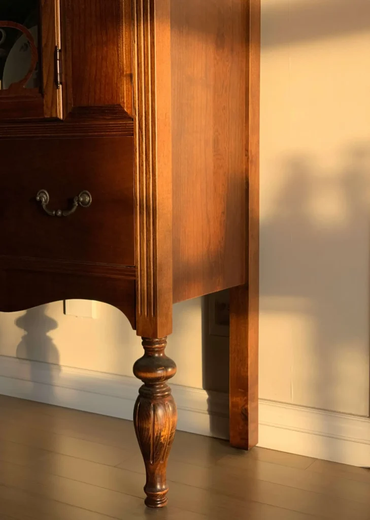 Close-up of a wooden cabinet with ornate leg and a decorative handle, bathed in warm sunlight, casting a shadow on the wall.
