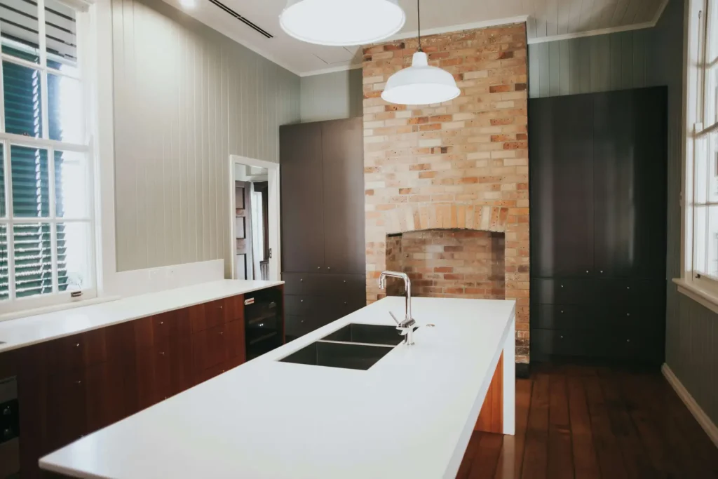 Modern kitchen with a white island countertop, sink, and overhead lights. Brick accent wall with a large archway and dark cabinetry.