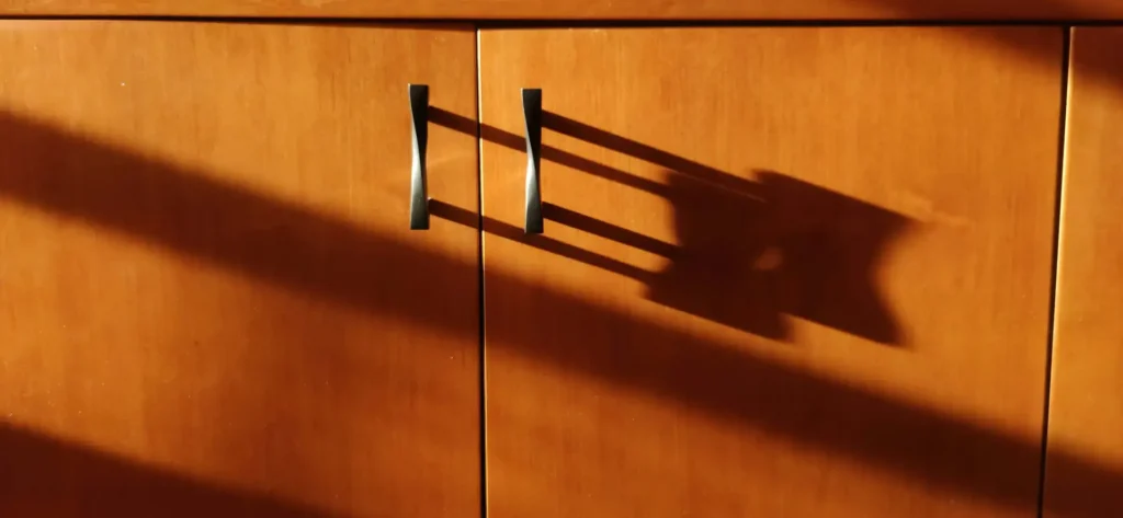 Wooden cabinet with two metal handles casting shadows, illuminated by sunlight.