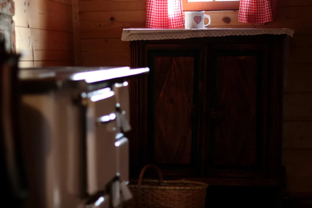 A cozy wooden room with a cabinet, a checkered curtain, and a mug on the windowsill. A woven basket sits on the floor beside an out-of-focus stove.