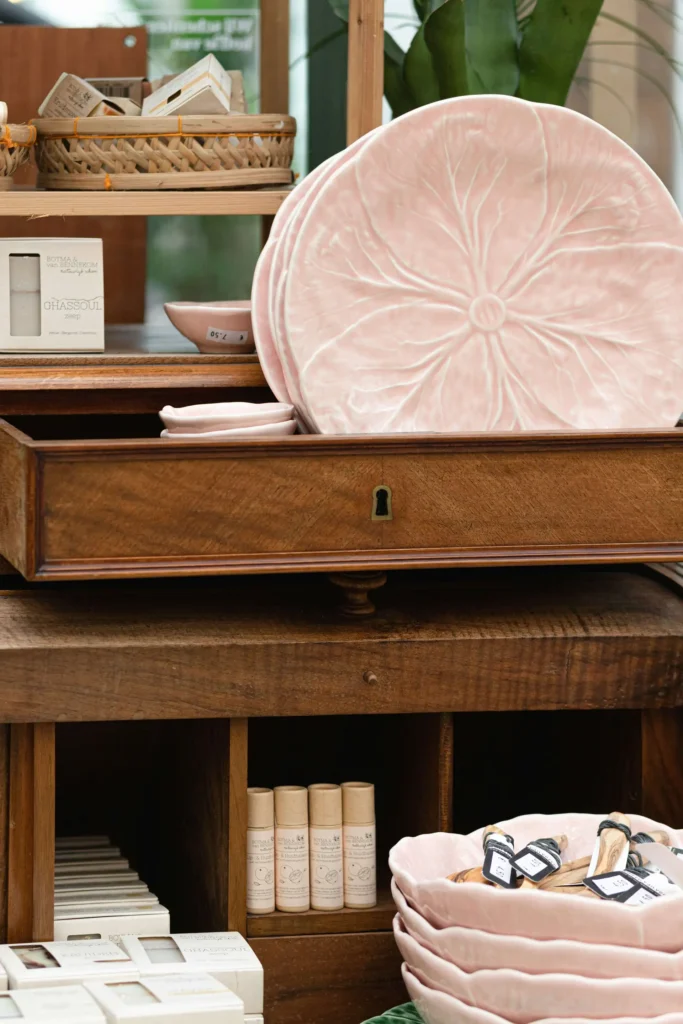 Wooden display with pink ceramic plates and various beauty products, including creams and lip balms, arranged on shelves and a drawer.