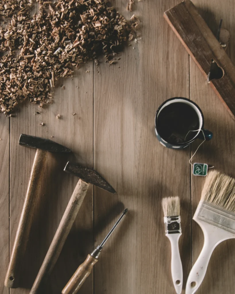 Wooden surface with sawdust, three hammers, a chisel, two paintbrushes, a teacup with a teabag, and a wooden plane.