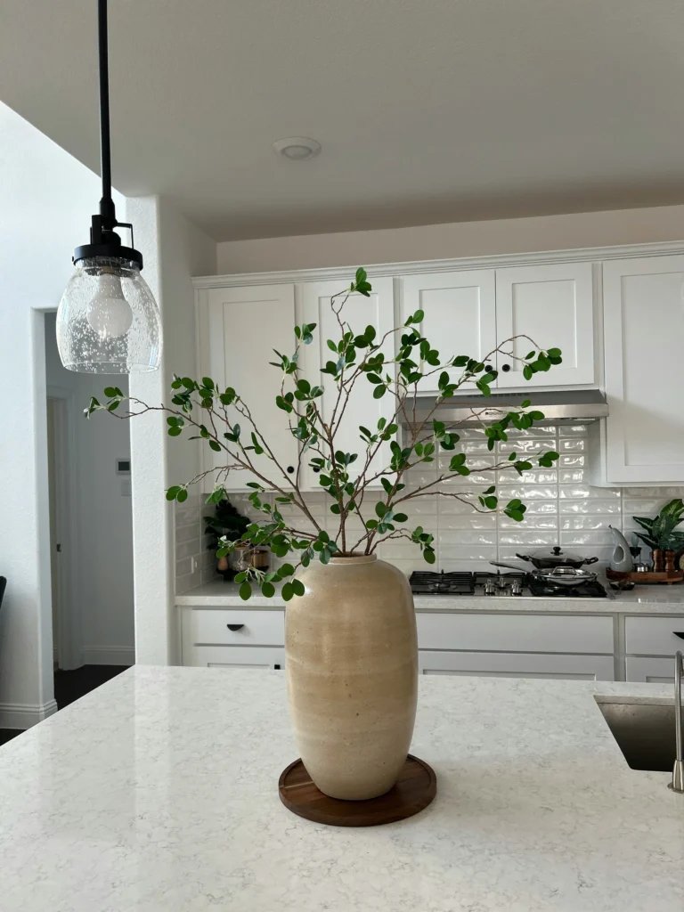 A minimalist kitchen features a large beige vase with green branches on a white island countertop, surrounded by white cabinets and a stove.