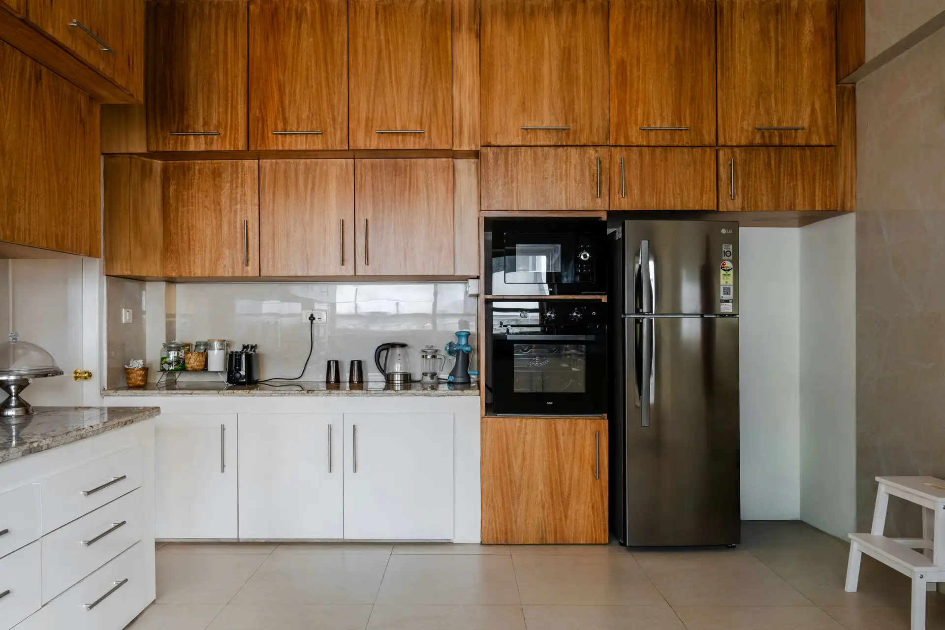 A modern kitchen with wooden cabinets, white drawers, a stainless steel fridge, built-in oven, and countertop appliances like a kettle and blender.
