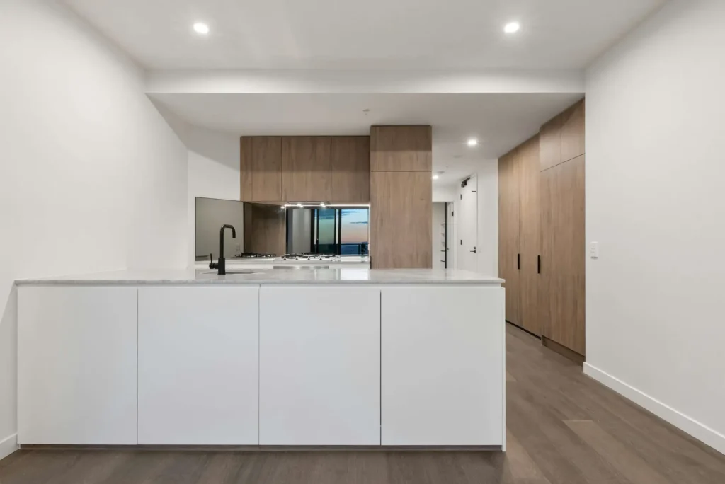 Modern kitchen with white cabinets, wood paneling, and a black faucet. There's a reflective backsplash and recessed ceiling lights, creating an open and minimalistic design.