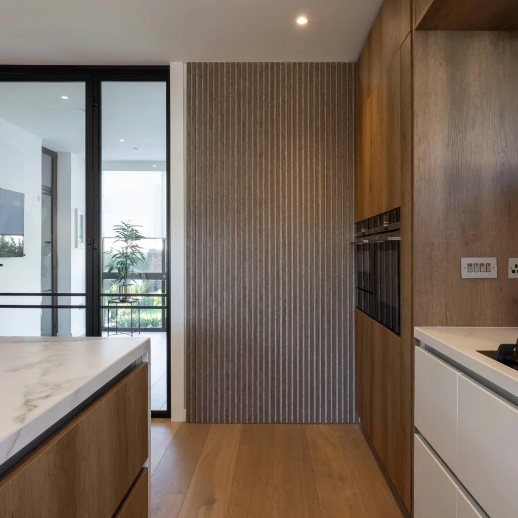 Modern kitchen with wooden cabinetry, marble countertop, and striped accent wall. Large glass door opens to a balcony with greenery visible outside.