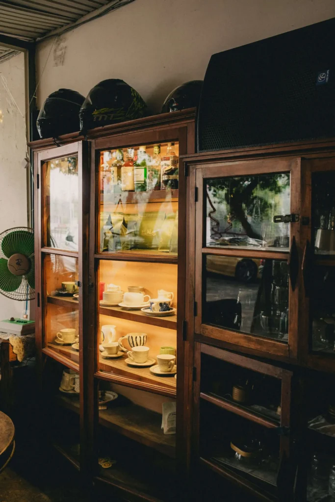 A wooden cabinet displaying various plates, cups, and bottles, with a small fan to the side and helmets on top.